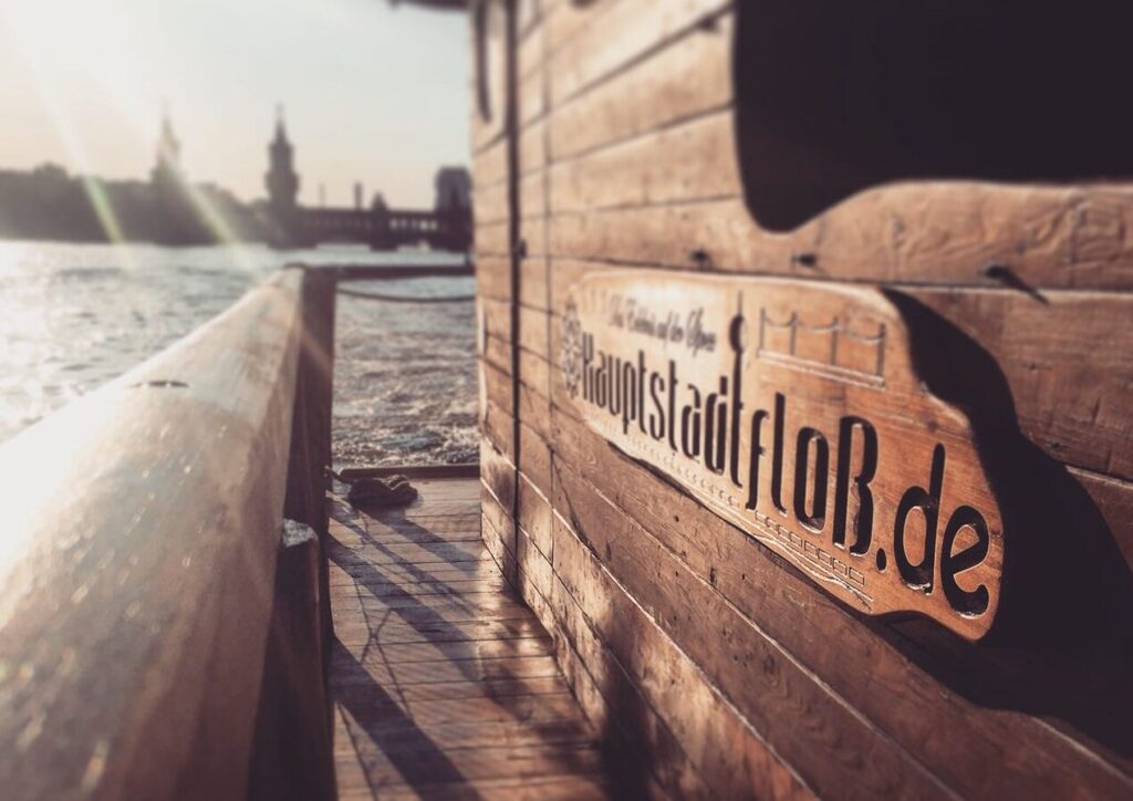 Hauptstadtfloß - Holzfloss am Ufer eines Flusses bei Sonnenuntergang, mit Blick auf eine Brücke und Türme im Hintergrund, eine idyllische Location für außergewöhnliche Orte zum Geburtstag feiern in Berlin.