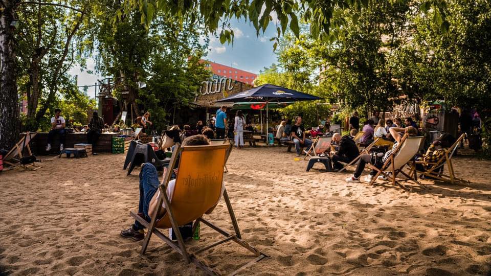 Entspannte Atmosphäre im YAAM Berlin mit Gästen, die auf Liegestühlen am Sandstrand verweilen.